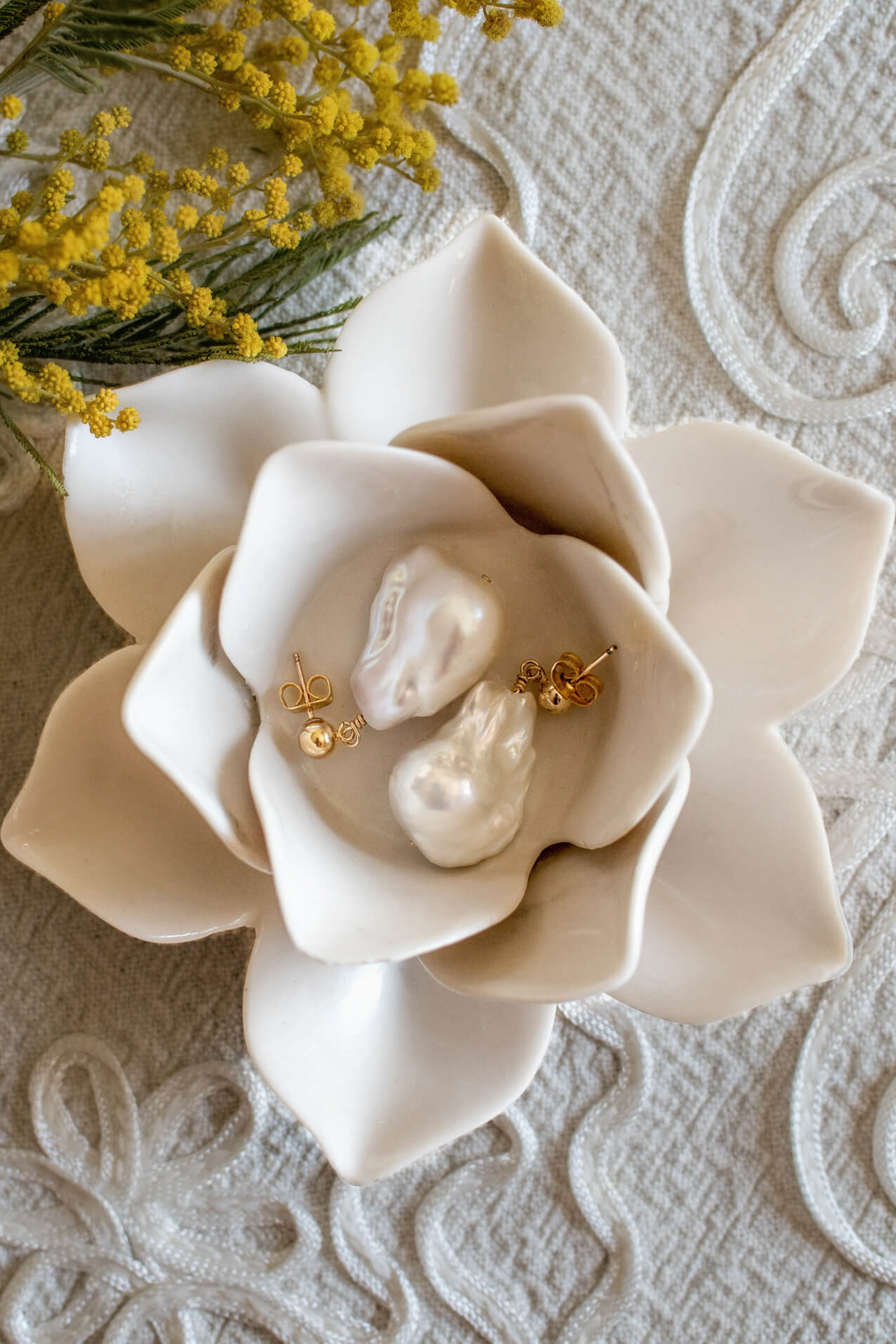 White ceramic flower-shaped dish with gold and pearl earrings on a textured surface.