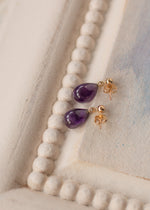 Amethyst teardrop earrings with gold hooks on a white beaded necklace.