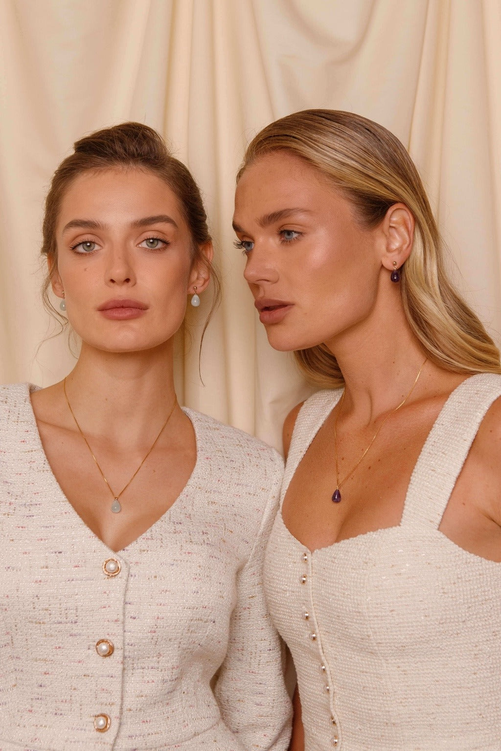 Two women wearing necklaces against a beige curtain background